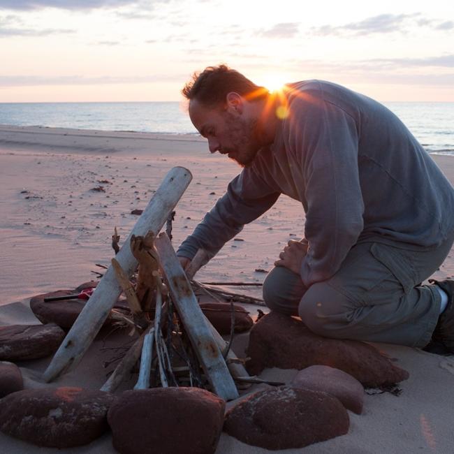 Royal Roads University professor Phillip Vanini kneels by a campfire built on a sandy beach.