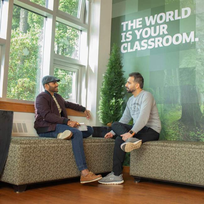 Two graduate students in conversation sit next to a window at Royal Roads University