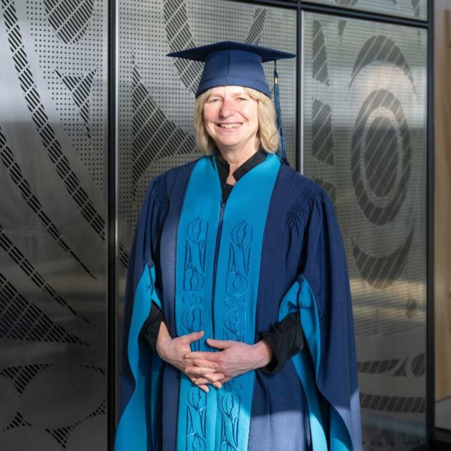 Suzanne Simard smiling in front of "All My Relations" installation wearing a cap and gown.