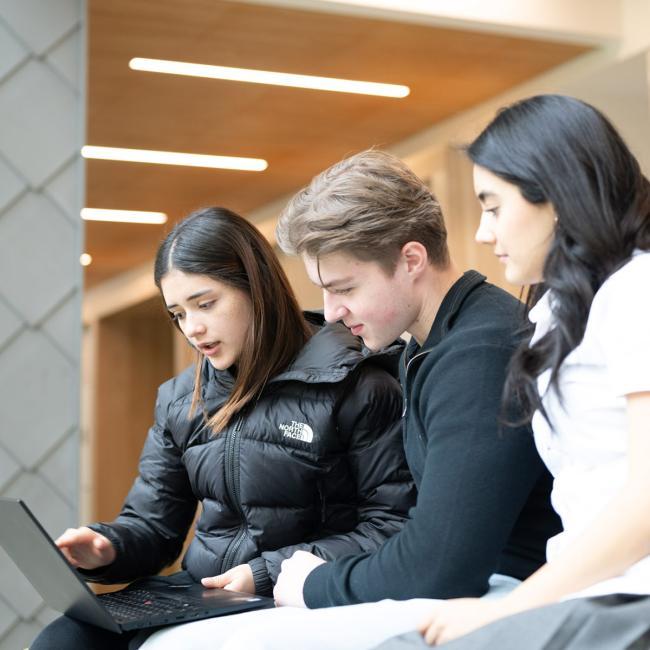 Three RRU undergraduate students look toward a laptop, sitting out front of a campus entrance
