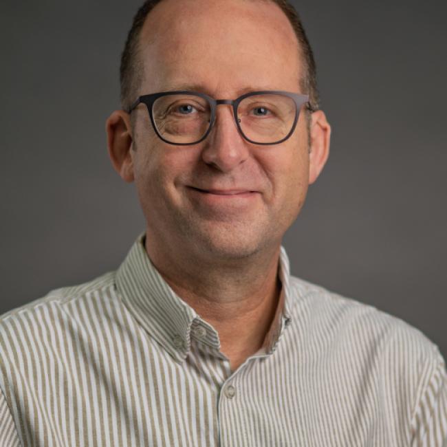 man with close cropped brown hair wearing glasses and a white collared shirt with black pinstripes. He's grinning slightly.
