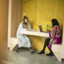 Two students sit in a booth with a yellow background in the Sherman Jen Building.