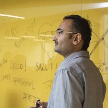 A smiling person person stands in front of a yellow marker board with greetings in different languages written all over it.