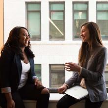 Two people sit in front of a large window in discussion, holding notebooks on their laps.