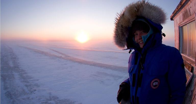 An icy sunset in Canada's far north, with snow visible on the ground. On the right side of the image is a woman in a large parka, the hood fully up, smiling into the cold.