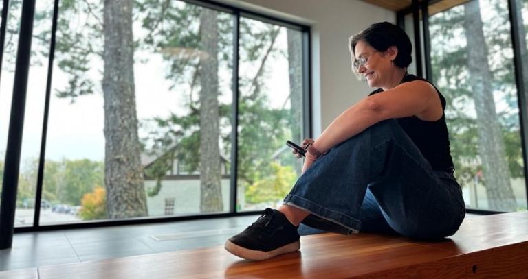 A woman scrolls on her phone in a lobby, trees can be seen out the window.